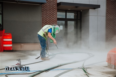  A worker in protective gear using a pressure washer to clean a concrete surface in front of a building, with orange safety barrels and hoses visible.