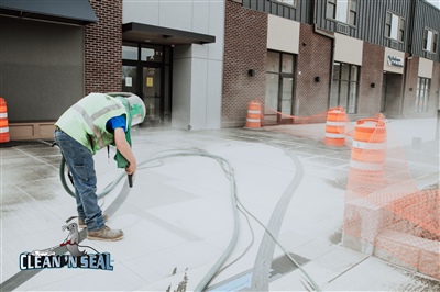 A worker in protective gear using a pressure washer to clean a concrete surface in front of a building, with orange safety barrels and hoses visible.