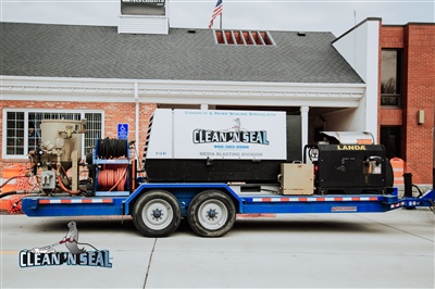 A Clean 'N Seal pressure washing trailer parked in front of a building, equipped with hoses and machinery.
