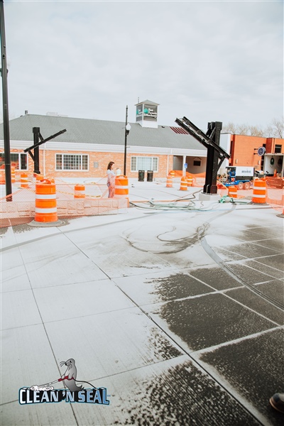 A newly pressure-washed concrete area with orange safety barrels and cones, surrounded by buildings and equipment.