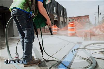 A worker in a green vest using a pressure washer on a concrete surface, with orange safety barrels and hoses visible, and buildings in the background.