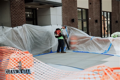 Two construction workers in safety vests working behind plastic sheeting at a building site, with orange safety netting in the foreground.
