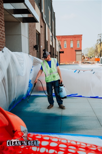 A construction worker in a yellow vest holding a paint can and roller, working on a site with plastic sheeting and orange safety netting.