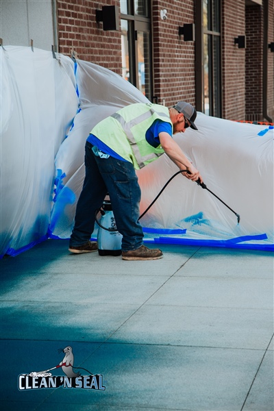  A construction worker in a yellow vest using a paint roller to apply blue paint on a concrete surface, with plastic sheeting in the background.