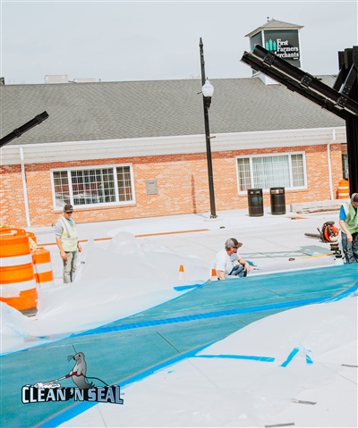 Construction workers applying blue paint to a concrete pathway in front of a brick building, with orange safety barrels and cones nearby.