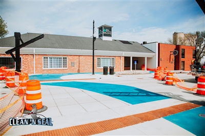 A newly painted blue and white concrete walkway in front of a brick building, surrounded by orange safety barrels and cones.