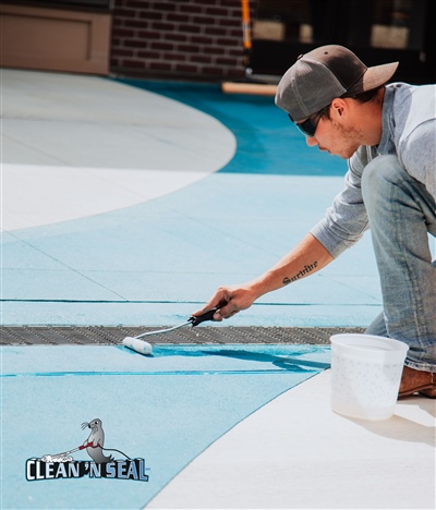 A worker in a cap and gray shirt using a roller brush to paint a concrete surface blue, with a white bucket nearby.