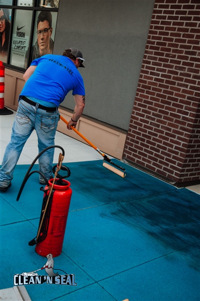 A worker in a blue shirt using a long-handled roller to apply a dark blue coating on a concrete surface, with a red fire extinguisher nearby.