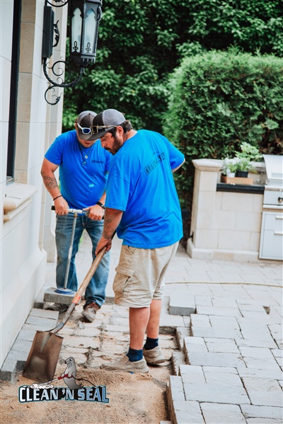 Two workers in blue shirts using shovels to dig and prepare a paved area next to a building, with greenery in the background.