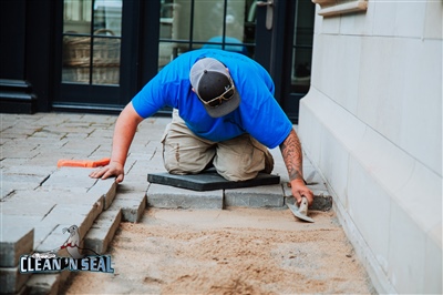 A worker in a blue shirt and cap kneeling on a padded mat, carefully placing paving stones next to a building wall.