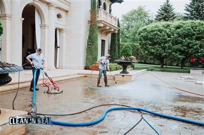  Two workers pressure washing the concrete driveway in front of an elegant building with arched windows and doors, surrounded by greenery.