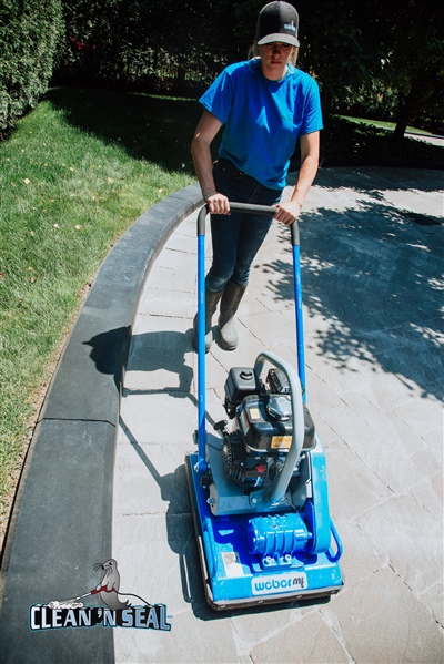  A worker in a blue shirt operating a blue power washer on a paved walkway next to a lawn area.