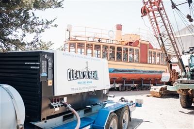  A heavy-duty pressure washing unit on a trailer in front of a large, wooden boat undergoing restoration, with cranes and other equipment in the background.