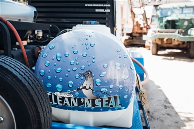 A close-up of a water tank on a pressure washing unit, featuring a graphic with water droplets and a seal, labeled "CLEAN 'N SEAL," with industrial equipment in the background.