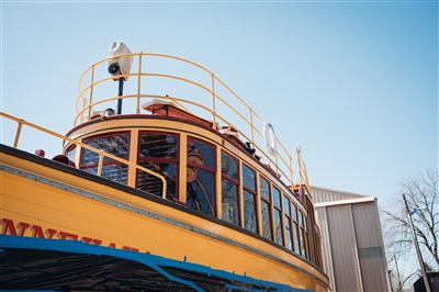  A close-up of the upper deck of a vintage wooden boat with yellow railings and a person visible inside, set against a clear blue sky.