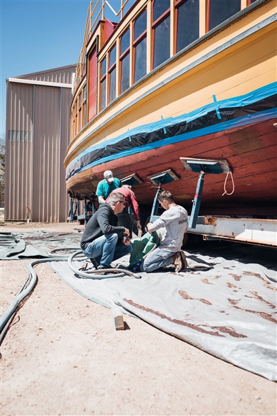  Workers kneeling and using tools to restore the underside of a vintage wooden boat, which is elevated on stands, with a building visible in the background.