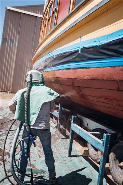 A worker in protective gear using a sandblaster to clean the underside of a vintage wooden boat, which is elevated on stands, with a building in the background.
