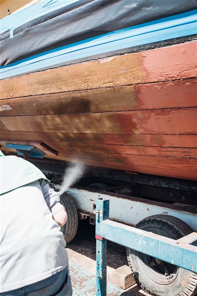  A worker in protective gear using a sandblaster to clean the wooden hull of a vintage boat, showing a contrast between the cleaned and uncleaned sections.