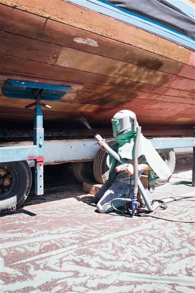 A worker in protective gear sitting on the ground, using a sandblaster to clean the wooden hull of a vintage boat, which is elevated on stands.