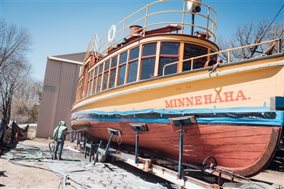 A vintage boat named "Minnehaha" elevated on stands, with a worker in protective gear sandblasting the wooden hull, and a building in the background.